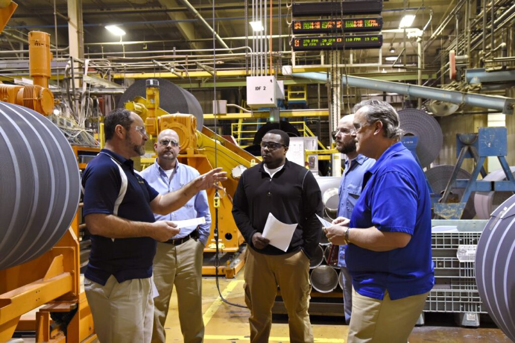 Five workers stand in the middle of a production facility in a circular formation, talking to each other.